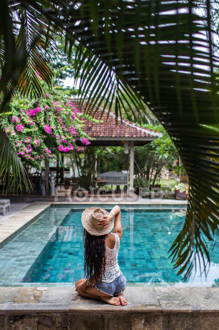 Giovane donna magra in pantaloncini jeans rilassante vicino piscina con cappello di paglia — Foto stock