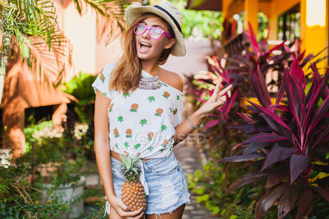 Attraente donna sorridente in vacanza in stampato t-shirt cappello di paglia moda estate, mani che tengono ananas — Foto stock