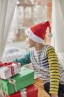 Menino em um chapéu de Papai Noel olhando pela janela . — Fotografia de Stock
