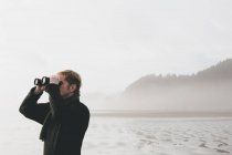 Hombre de pie en la playa y mirando a través de prismáticos - foto de stock