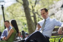 Человек в костюме смотрит на смартфон, сидя на скамейках в парке с коллегами — стоковое фото