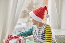 Garçon dans le chapeau de Père Noël regardant par la fenêtre avec cadeau de Noël de seuil de fenêtre . — Photo de stock