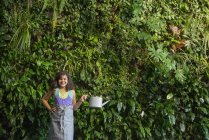 Elementary age girl standing in front of wall covered with ferns and climbing plants with watering can. — Stock Photo