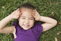 Mädchen im Grundalter liegt mit den Händen am Kopf im Gras und lacht. — Stockfoto