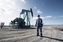 Man in overalls and hard hat at pump jack in open ground at oil extraction site. — Stock Photo