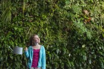 Девушка младшего возраста, стоящая перед стеной, покрытая папоротниками и растениями для скалолазания с помощью лейки . — стоковое фото