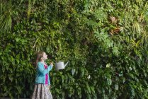 Mädchen im Grundschulalter vor Wand mit Farnen und Kletterpflanzen mit Gießkanne. — Stockfoto