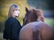 Young woman leading bay horse by bridle and looking in camera. — Stock Photo