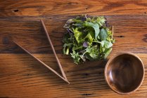 Round polished wooden bowl and clutch of organic mixed salad leaves with wooden chopsticks. — Stock Photo