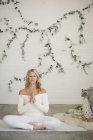 Блондинка сидить на білому килимку йоги в положенні лотоса з затиснутими руками . — стокове фото