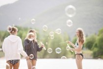 Adolescentes de pie junto al lago rodeado de burbujas de jabón al aire libre . - foto de stock