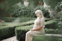 Vista lateral da mulher grávida em vestido branco sentado no jardim verde . — Fotografia de Stock
