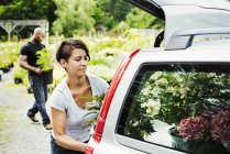Femme chargeant des fleurs dans le coffre de la voiture garée au centre de jardin avec l'homme portant des plantes en arrière-plan . — Photo de stock