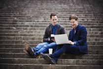 Два молодых человека сидят на ступенях города и смотрят на ноутбук вместе . — стоковое фото