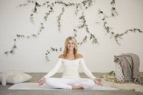 Блондинка сидит на белом коврике для йоги и медитирует. . — стоковое фото