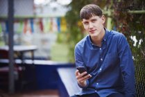Young man sitting in city, holding smartphone and looking in camera. — Stock Photo