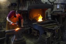 Herrero hembra golpeando metal rojo caliente en yunque dentro del taller . - foto de stock