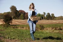 Mid adult woman walking across field and holding basket of crops. — Stock Photo