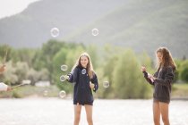 Ragazze adolescenti in piedi vicino al lago circondate da bolle di sapone all'aperto . — Foto stock