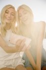 Duas meninas adolescentes loiras tirando selfie com smartphone contra luz suave . — Fotografia de Stock
