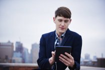 Young businessman with earphones standing on rooftop and using digital tablet. — Stock Photo