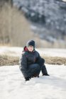 Boy in winter coat and woolly hat crouching in snow. — Stock Photo
