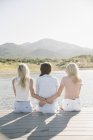 Rear view of mother and blonde daughters sitting on lake jetty. — Stock Photo