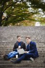 Двоє молодих чоловіків сидять на сходах парку і використовують ноутбук разом . — стокове фото