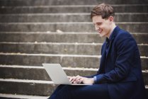 Jovem sentado em degraus na cidade e trabalhando no laptop . — Fotografia de Stock