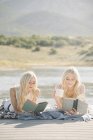 Due ragazze adolescenti bionde che leggono libri sul molo del lago . — Foto stock