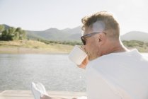 Homem sentado no lago molhe e bebendo de caneca de café . — Fotografia de Stock