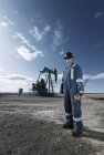 Man in overalls and hard hat at pump jack in open ground at oil extraction site. — Stock Photo