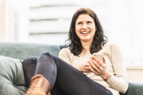 Frau sitzt mit den Füßen auf Sofa, lacht und hält Smartphone. — Stockfoto