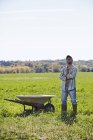 Homem adulto médio com braços cruzados em pé no campo de cultivo ao lado do carrinho de mão . — Fotografia de Stock