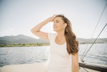 Mulher com cabelos castanhos longos em pé no veleiro e ajustando óculos de sol no lago . — Fotografia de Stock