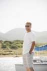 Blond man wearing sunglasses and standing on lake jetty by sailboat. — Stock Photo