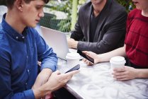 Tres personas que trabajan en la mesa al aire libre al aire libre con teléfonos inteligentes y portátil . - foto de stock