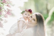 Mid adult woman posing with baby daughter with flower wreath outdoors. — Stock Photo