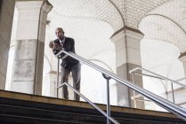 Maduro hombre de negocios en traje de pie junto a la escalera bajo el techo arqueado y la celebración de teléfonos inteligentes . - foto de stock
