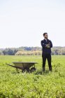 Young man with arms crossed standing in crop field next to wheelbarrow. — Stock Photo