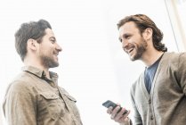 Два бизнесмена улыбаются и держат смартфон в офисе . — стоковое фото