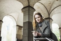 Woman with long hair holding smartphone and looking in camera under archway. — Stock Photo