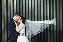 Bride with windblown veil and bridegroom kissing in front of green corrugated metal wall, side view. — Stock Photo