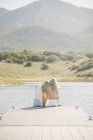 Visão traseira de duas meninas loiras sentadas no molhe do lago . — Fotografia de Stock