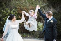 Bride and bridegroom couple holding and swinging girl in air outdoors. — Stock Photo