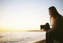 Вид збоку жінки з білявим волоссям, що сидить на піщаному пляжі і тримає камеру . — стокове фото
