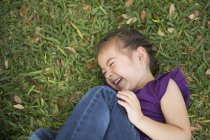 Primer plano de la niña acostada, abrazando las rodillas y riendo en la hierba verde . - foto de stock