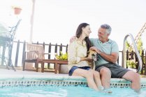 Seniorenpaar sitzt am Rand des Swimmingpools und streichelt Hund dazwischen. — Stockfoto