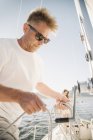 Portrait of blond man with sunglasses holding ropes on sailboat. — Stock Photo