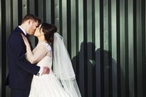 Noiva e noivo beijando na frente da parede de metal ondulado verde, vista lateral . — Fotografia de Stock